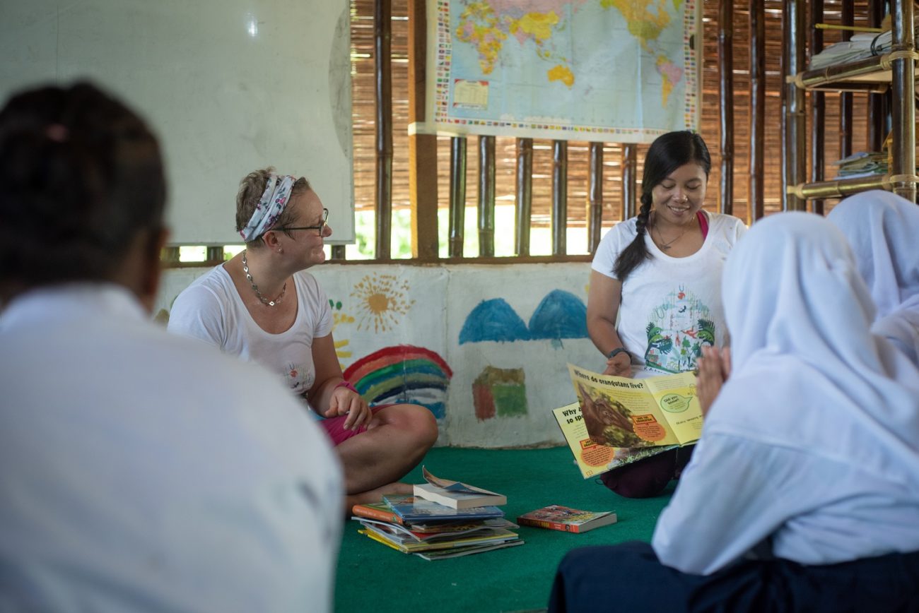 Two women sitting at the front of a classroom. One is reading a book to the class.