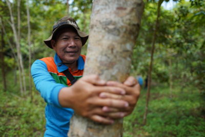 A man is standing with his hands wrapped around a tree trunk. He is smiling into the camera.