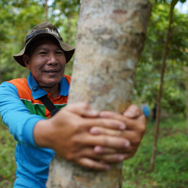 A man is standing with his hands wrapped around a tree trunk. He is smiling into the camera.