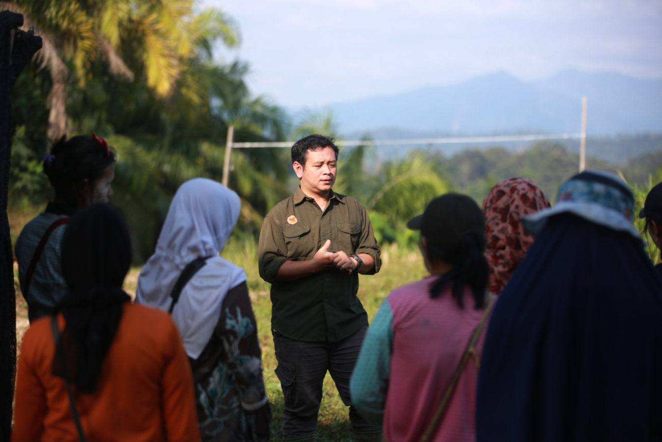 A man in uniform is talking to a group of people. They are outside and there is forest in the background.