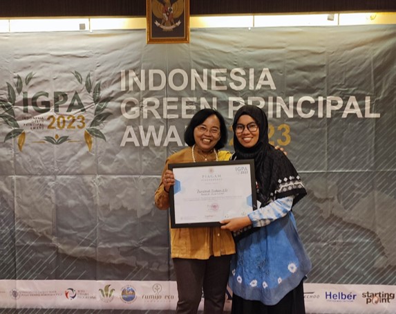 Two smiling women stand in front of a banner reading 'Indonesia Green Principal Award 2023', one is handing the other a framed award.
