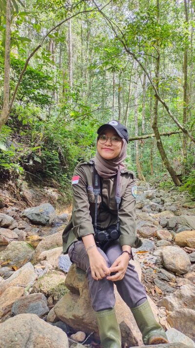 A woman - Wita Azanraini Fadillah- Batang Toru Village Facilitator - sits on a rock in a dried river bed in a forest