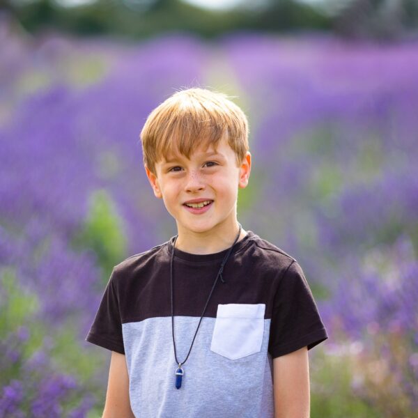 A young boy stands against a field of lavender, smiling towards the camera