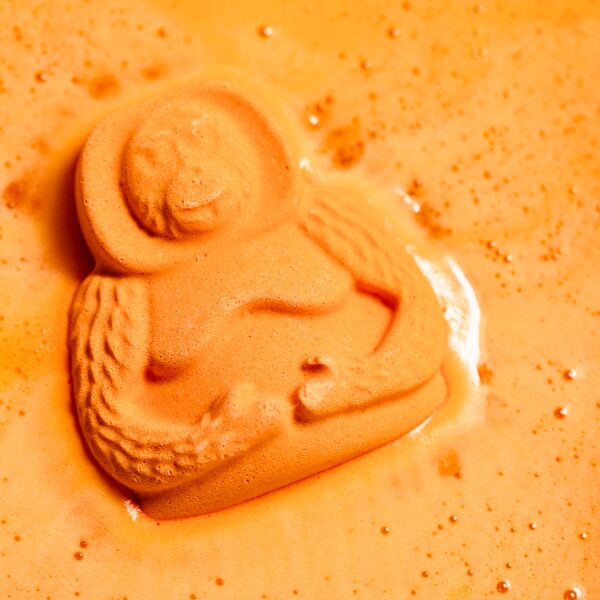 A bath bomb shaped like an orangutan fizzes in water, leaving orange foam behind