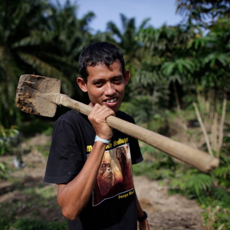 Krisna holds a shovel over his shoulder. His t-shirt has an orangutan on it.