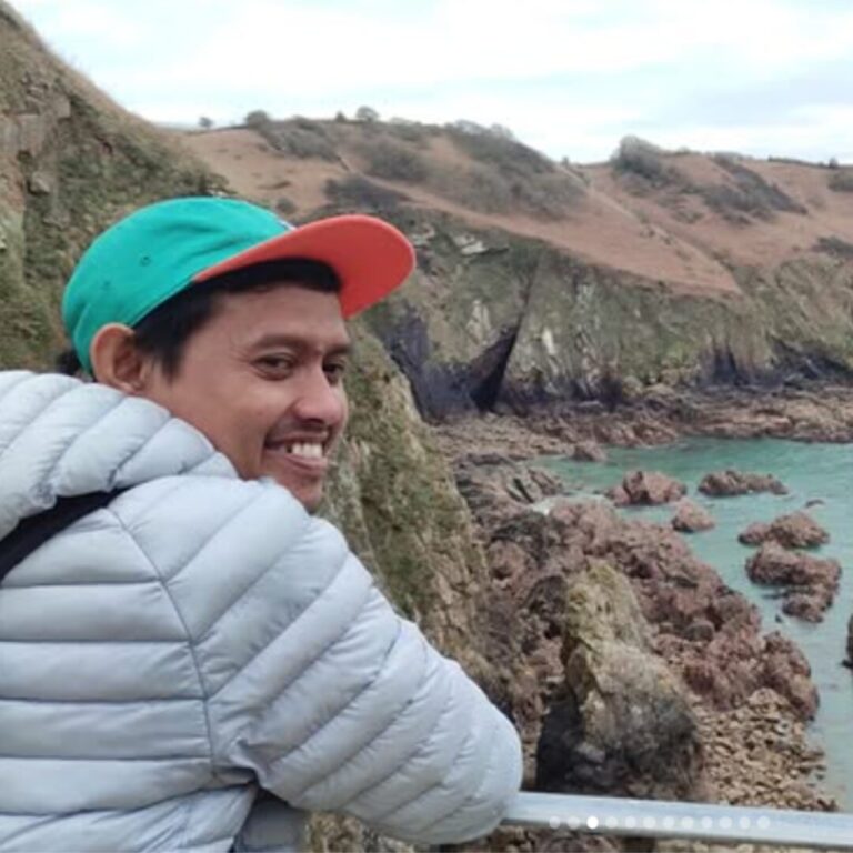 Krisna looks over his shoulder and smiles. The background shows the cliffs and sea in Jersey.